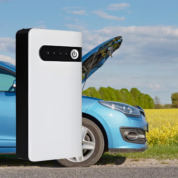 Emergency car battery jump starter kit shown in front of a broken-down car with open hood.