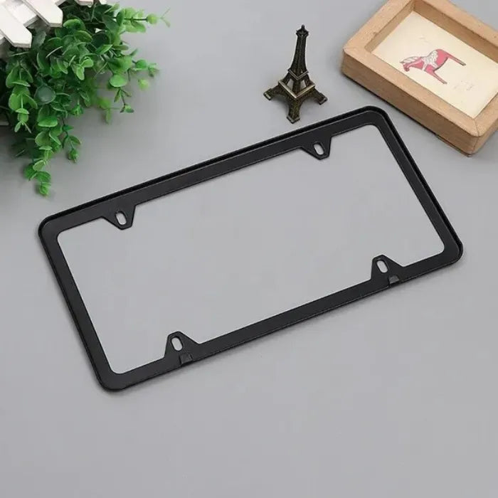 Black license plate holder displayed with mini Eiffel Tower and decor on flat gray surface.