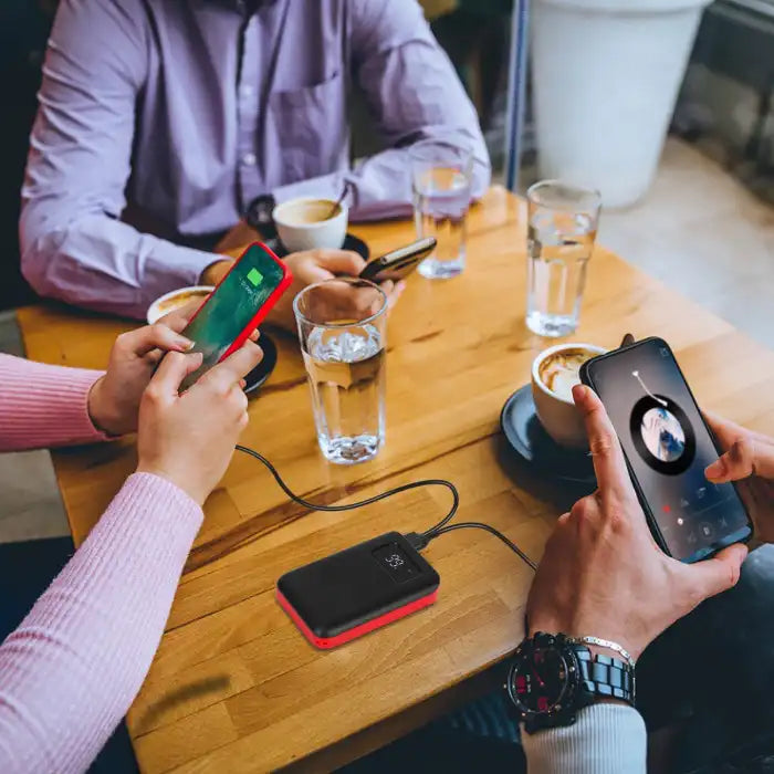 Power Bank charging two smartphones at a café table with people using their devices during coffee time.